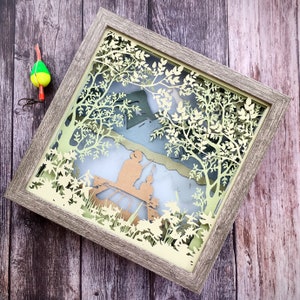 Op de afbeelding: Een ingelijst papieren kunstwerk met een 3D-effect. De kunst toont een gedetailleerd uitgesneden ontwerp van bomen en bladeren, met een silhouet van twee mensen op de achtergrond. De lijst is van lichtbruin hout.