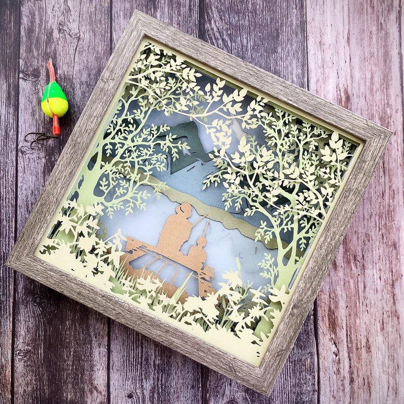 Op de afbeelding: Een ingelijst papieren kunstwerk met een 3D-effect. De kunst toont een gedetailleerd uitgesneden ontwerp van bomen en bladeren, met een silhouet van twee mensen op de achtergrond. De lijst is van lichtbruin hout.
