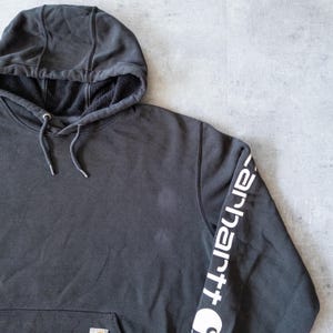 Puede incluir: Una sudadera con capucha gris carb&oacute;n con capucha con cord&oacute;n y bolsillo tipo canguro. La manga izquierda presenta el logotipo blanco de Carhartt. El interior de la capucha est&aacute; forrado con forro polar negro. Prenda casual y c&oacute;moda.