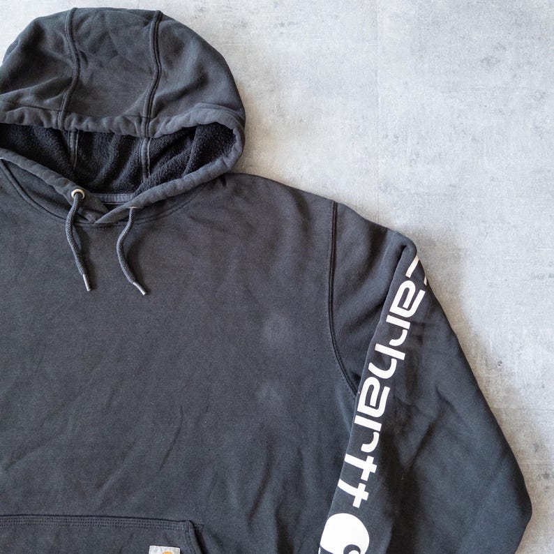 Puede incluir: Una sudadera con capucha gris carb&oacute;n con capucha con cord&oacute;n y bolsillo tipo canguro. La manga izquierda presenta el logotipo blanco de Carhartt. El interior de la capucha est&aacute; forrado con forro polar negro. Prenda casual y c&oacute;moda.