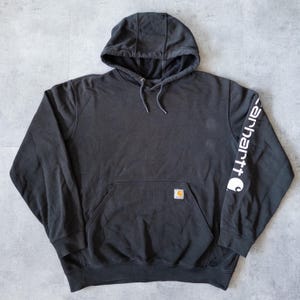 Puede incluir: Una sudadera con capucha negra con un bolsillo de canguro y el nombre de la marca "Carhartt" impreso en blanco en la manga izquierda. La sudadera tiene una capucha con cord&oacute;n y un peque&ntilde;o parche con el logotipo.