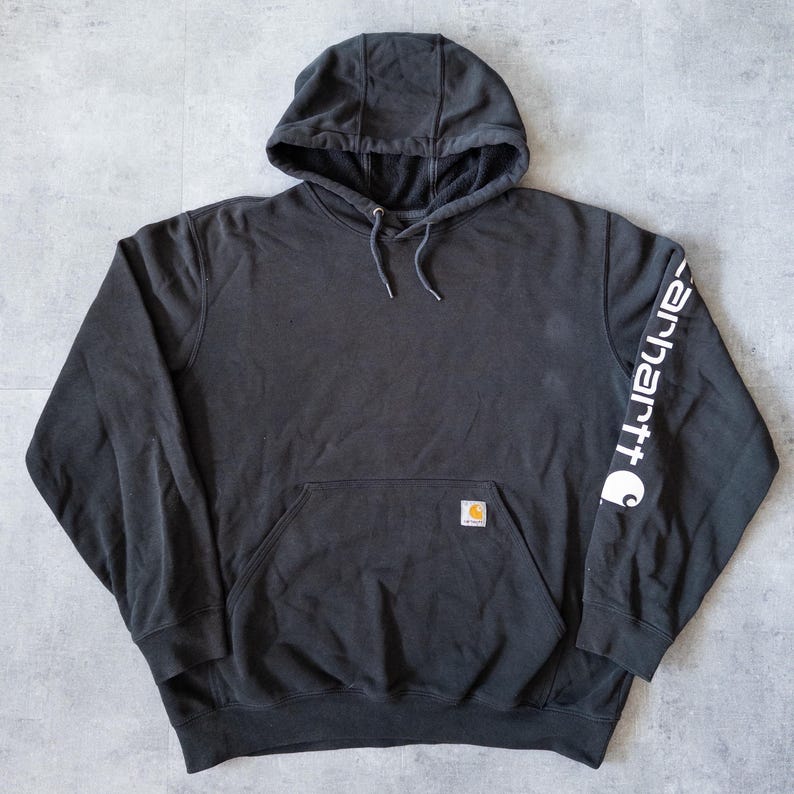 Puede incluir: Una sudadera con capucha negra con un bolsillo de canguro y el nombre de la marca "Carhartt" impreso en blanco en la manga izquierda. La sudadera tiene una capucha con cord&oacute;n y un peque&ntilde;o parche con el logotipo.