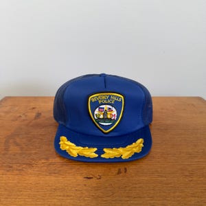 Pode incluir: Boné azul de caminhoneiro com parte traseira em rede e aba curva. A frente apresenta um patch com as palavras "Beverly Hills Police" e um emblema dourado. Detalhes de folhas douradas são bordados na aba.