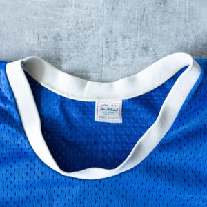 May include: Close-up of a vintage blue mesh jersey with a white collar and trim. The jersey has a tag that reads "LARGE 42-44" and "Don Allen". The jersey is made of 100% nylon.