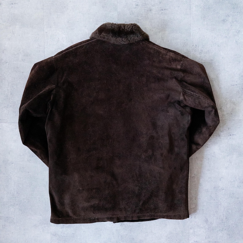 K&ouml;nnte beinhalten: Dunkelbraune Wildlederjacke mit Kunstfellkragen. Die Jacke hat lange &Auml;rmel und einen Knopfverschluss. Die R&uuml;ckseite der Jacke ist sichtbar und zeigt die Textur des Wildledermaterials. Die Jacke ist ein klassischer Stil.