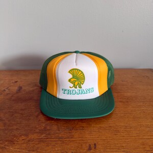 May include: A green, yellow, and white trucker hat with the word "TROJANS" in green. The hat features a green brim and mesh back, with a yellow and white front panel. The front panel displays a green graphic of a helmet.