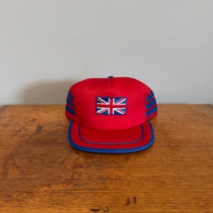 Op de afbeelding: Een felrode honkbalpet met een centraal geborduurde patch van de Union Jack-vlag. De pet heeft een gestructureerd voorpaneel, een platte klep met blauwe rand en blauwe mesh zijpanelen met drie horizontale blauwe strepen aan elke kant.