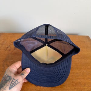May include: Navy blue trucker hat with a mesh back and a curved peak. The hat has a structured front panel and an adjustable snap closure. The interior has a white lining. The hat is designed for casual wear and sun protection.