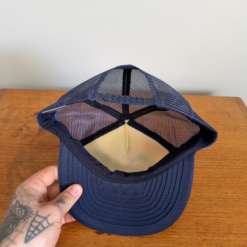 May include: Navy blue trucker hat with a mesh back and a curved peak. The hat has a structured front panel and an adjustable snap closure. The interior has a white lining. The hat is designed for casual wear and sun protection.