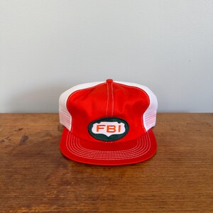 May include: A red and white trucker hat featuring a white oval patch with a dark green border and the letters FBI in orange. The cap has a structured red front panel, a flat red brim with white stitching, and white mesh side and back panels.
