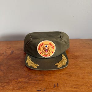 May include: Olive green military-style cap with a circular patch featuring the United States Army emblem. The cap has gold embroidered accents on the brim. The patch is in red, white, and blue.