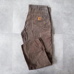 Puede incluir: Pantalones de trabajo marrones con m&uacute;ltiples bolsillos y un parche con el logo de Carhartt en el bolsillo trasero. Los pantalones est&aacute;n hechos de un material duradero y tienen un ajuste relajado. Los pantalones son talla 42.