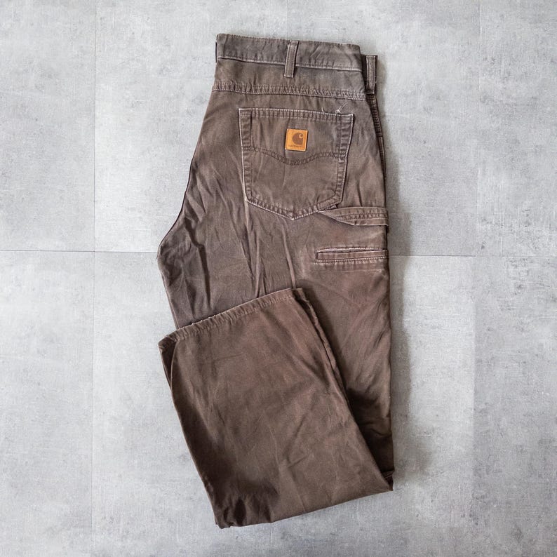 Puede incluir: Pantalones de trabajo marrones con m&uacute;ltiples bolsillos y un parche con el logo de Carhartt en el bolsillo trasero. Los pantalones est&aacute;n hechos de un material duradero y tienen un ajuste relajado. Los pantalones son talla 42.