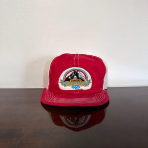 May include: A red trucker hat with white mesh side and back panels. The front features a white patch with a colorful rainbow arch over a black mountain graphic and the text Canada's Wonderland in blue and yellow lettering.