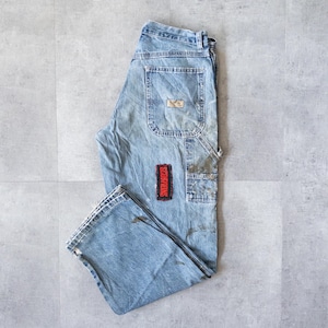 May include: Faded light blue denim work trousers with multiple pockets. A red patch with the word "SCORPIONS" is sewn on the front. The trousers have a relaxed fit and show signs of wear and tear.