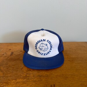 Puede incluir: Gorra de camionero azul y blanca con visera curva. La gorra muestra el texto "Georgian College Grizzlies" en un diseño circular en la parte delantera. Tiene una parte trasera de malla y cierre a presión.