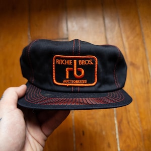 Op de afbeelding: Zwarte pet met een platte klep en rode stiksels. De voorkant heeft een rechthoekige patch met de woorden "Ritchie Bros." en "Auctioneers" in oranje. De patch toont ook de letters "rb" in oranje.