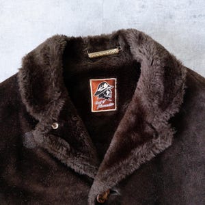 K&ouml;nnte beinhalten: Braune Wildlederjacke mit Kunstfellkragen. Die Jacke hat einen Knopfverschluss und ein Etikett mit Logo und Text. Die Jacke ist im Vintage-Stil.