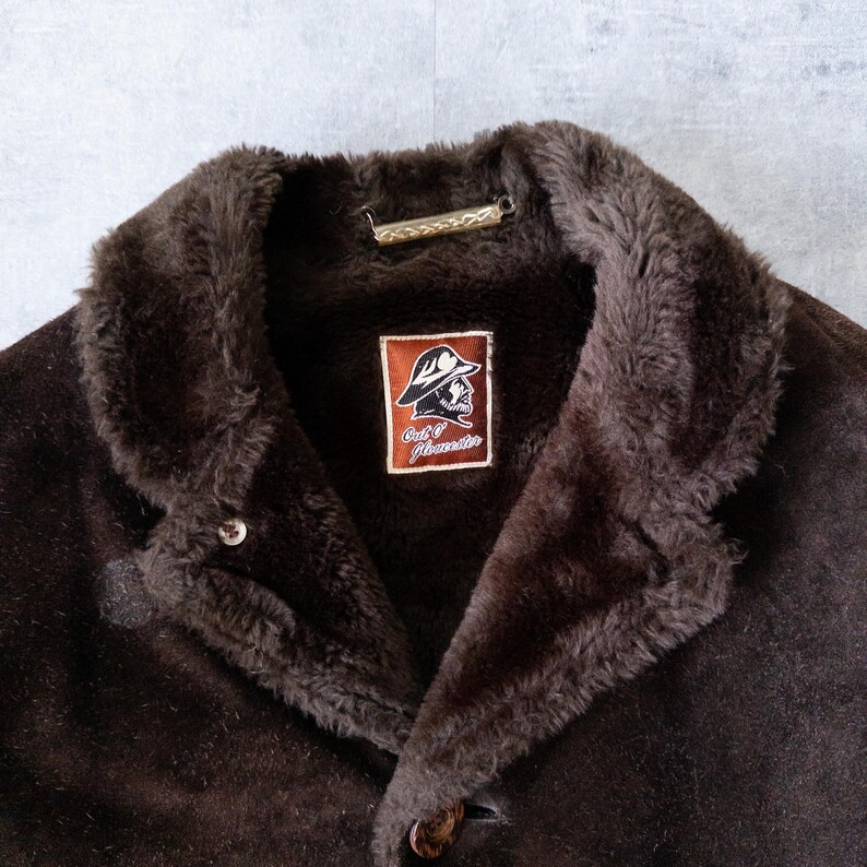 K&ouml;nnte beinhalten: Braune Wildlederjacke mit Kunstfellkragen. Die Jacke hat einen Knopfverschluss und ein Etikett mit Logo und Text. Die Jacke ist im Vintage-Stil.