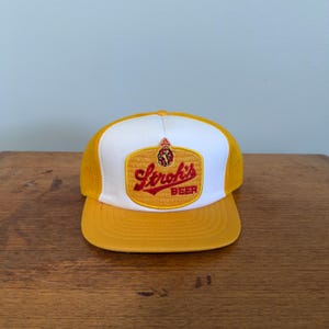 May include: A yellow and white trucker hat featuring the Stroh's Beer logo. The hat has a yellow peak and mesh back, with a white front panel. The logo is a gold rectangle with red lettering. A classic cap.