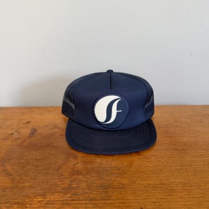 May include: A navy blue trucker hat featuring a solid front panel with a circular white patch displaying a stylized letter F. The cap includes a structured crown, a flat brim, and mesh side and back panels for ventilation, finished with a button top.