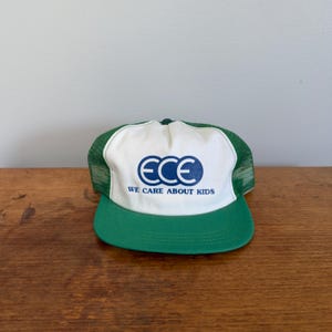 May include: A green and white trucker cap featuring a white front panel with the blue text ECE and the phrase WE CARE ABOUT KIDS printed below. The cap has a green brim and green mesh side and back panels with a button on the top centre.