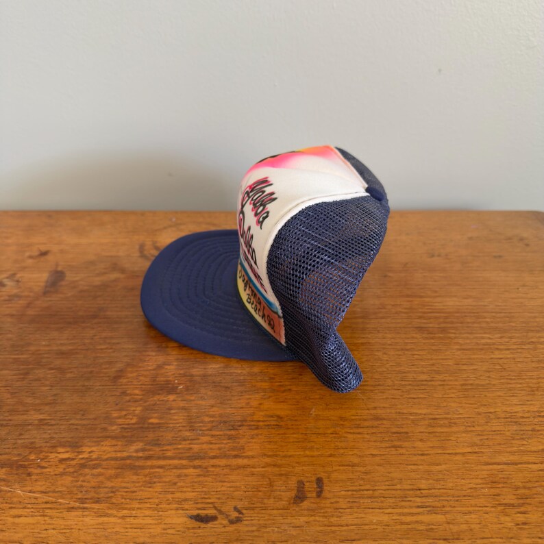 May include: A navy blue trucker hat with a white front panel displaying the word "Aloha". The hat has a mesh back and a curved brim. The front panel features a colourful design with pink, orange, and yellow hues.