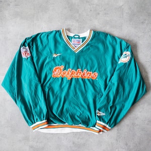 Puede incluir: Chaqueta cortavientos vintage de los Dolphins de color verde azulado con detalles en naranja y blanco. Presenta el logotipo de los Dolphins, el logotipo de la NFL y el logotipo de Reebok. La palabra "Dolphins" está escrita en naranja en el pecho.