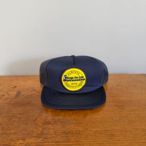 Op de afbeelding: Een marineblauwe truckerpet met een ronde gele patch op de voorkant. De patch bevat de tekst Alberta Change the Law Campaign Federation of Labour in zwarte letters. De pet heeft een stevig voorpaneel en een achterkant van gaas.