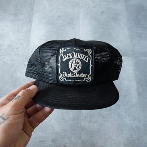 Puede incluir: Gorra de camionero negra con parte trasera de malla y visera curva. La parte delantera presenta un parche rectangular con el logotipo de Jack Daniel's en letras blancas y borde negro. Un accesorio clásico.