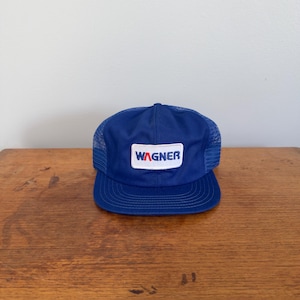 May include: A royal blue trucker hat featuring a mesh back and a solid front panel. A rectangular white patch is centered on the front, displaying the word WAGNER in blue capital letters with a red letter A, stitched in a bold, sans-serif font.
