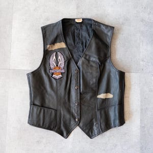 Peut inclure: Gilet en cuir noir avec un col en V et une fermeture boutonnée. Comprend un écusson Harley-Davidson avec un emblème ailé et le texte "MOTOR HARLEY-DAVIDSON CYCLES". Avec deux poches avant et une petite étiquette.