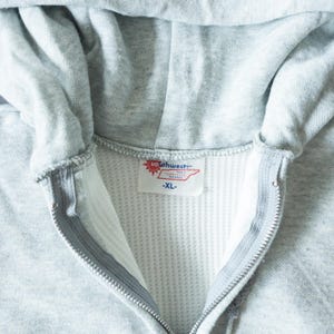 Pu&ograve; includere: Felpa con cappuccio grigio chiaro con cerniera, cappuccio e fodera a nido d'ape bianca. La cerniera &egrave; argentata. L'etichetta recita "southwest" e "-XL-".