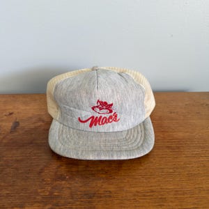 Puede incluir: Gorra de camionero gris con visera curva y parte trasera de malla. La parte delantera de la gorra presenta el texto rojo "Mac's" con un logotipo de gato de dibujos animados rojo. La gorra está sobre una superficie de madera.