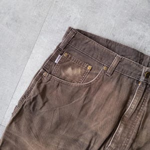 Puede incluir: Primer plano de pantalones de trabajo marrones con desgaste visible. Los pantalones presentan un dise&ntilde;o cl&aacute;sico de cinco bolsillos, remaches met&aacute;licos y una peque&ntilde;a etiqueta de marca. La tela tiene un aspecto descolorido.