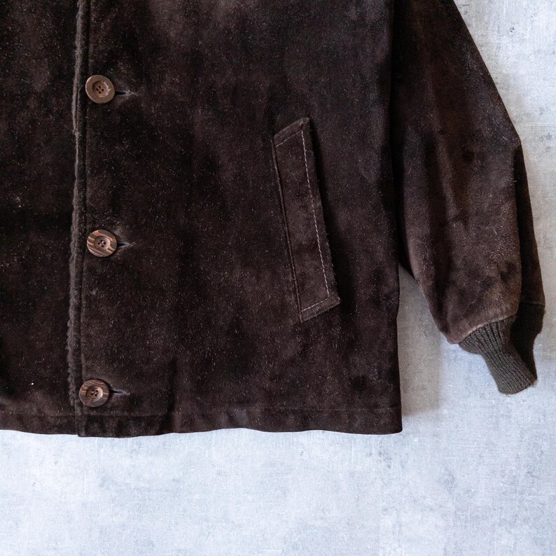K&ouml;nnte beinhalten: Nahaufnahme einer braunen Wildlederjacke mit drei Kn&ouml;pfen und einer Tasche. Die Jacke hat eine gerippte Manschette am &Auml;rmel. Das Wildleder hat ein leicht abgenutztes Aussehen.