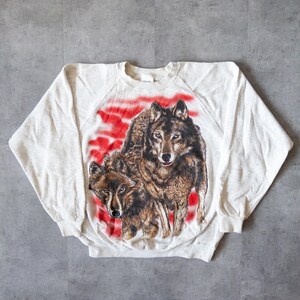 Könnte beinhalten: Ein hellgraues Sweatshirt mit einer detaillierten Grafik von zwei Wölfen. Die Wölfe sind in Braun- und Schwarztönen gehalten und vor einem rot-weißen Hintergrund platziert. Das Sweatshirt hat lange Ärmel und einen Rundhalsausschnitt.