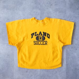 May include: A mustard yellow short-sleeved sweatshirt featuring the navy blue text "PLANO 13 SOCCER". The shirt has a crew neck and is made of a soft material.