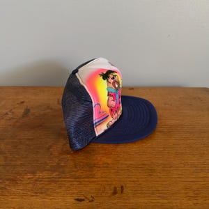 May include: A navy blue trucker hat with a cartoon character illustration on the front. The character is wearing a pink and blue outfit, set against a yellow and pink backdrop. The hat has a white front panel, a navy blue brim, and a mesh back.