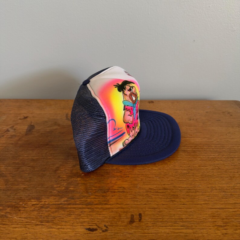 May include: A navy blue trucker hat with a cartoon character illustration on the front. The character is wearing a pink and blue outfit, set against a yellow and pink backdrop. The hat has a white front panel, a navy blue brim, and a mesh back.