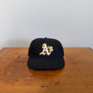 May include: Black baseball cap featuring the Oakland Athletics team logo. The logo is a stylized "A's" in white with a gold outline. The cap has a curved brim and a structured crown.