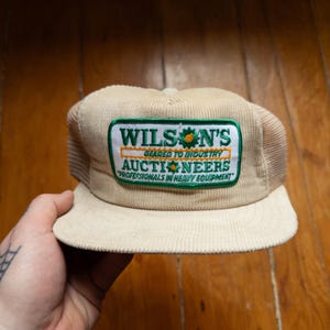 Może przedstawiać: Beżowa czapka typu trucker z sztruksu z siatkowym tyłem. Na przedniej naszywce widnieje napis "WILSON'S AUCTIONEERS" w kolorze zielono-białym, z tekstem "GEARED TO INDUSTRY" i "PROFESSIONALS IN HEAVY EQUIPMENT".