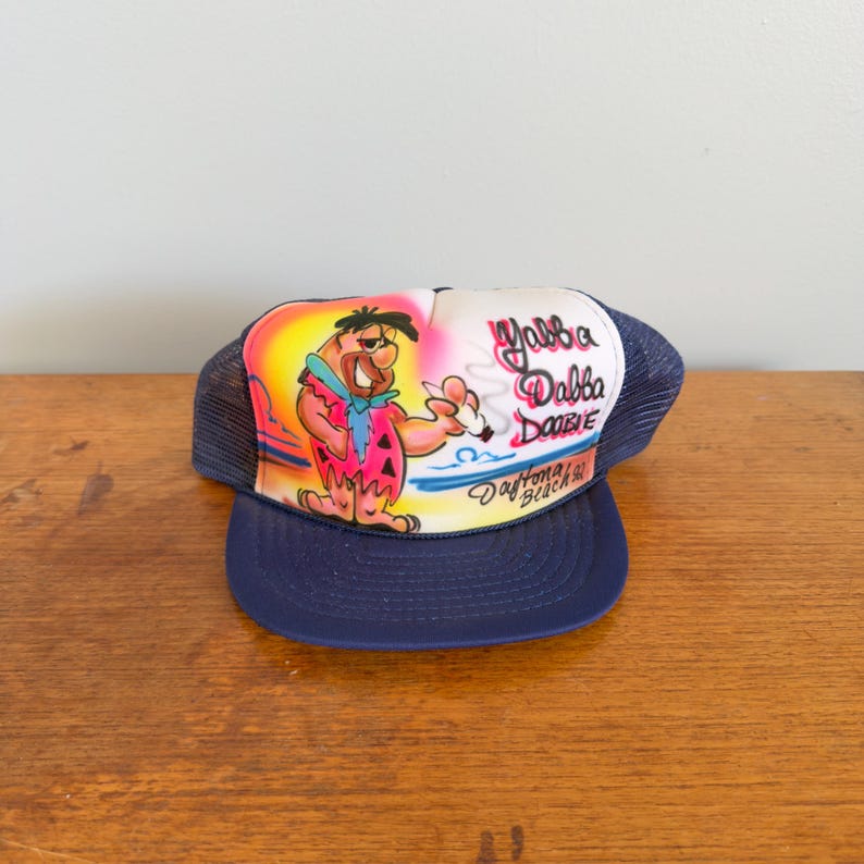 May include: A navy blue trucker hat with a cartoon image of Fred Flintstone, the words "Yabba Dabba Doo" and "Daytona Beach" printed on the front. The hat has a mesh back and a curved peak.