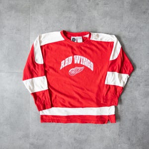 May include: A red long-sleeved shirt with white panels on the shoulders and sleeves. The shirt displays the words "RED WINGS" in white arched lettering above a white logo of a winged wheel. The shirt is made of a soft material.