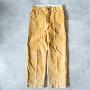 Op de afbeelding: Mosterdkleurige corduroy broek met een knoopsluiting en zakken. De broek heeft een rechte pijp en een vervaagde uitstraling. Het kledingstuk ligt plat op een grijze ondergrond en toont zijn textuur en kleur.