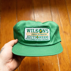 Könnte beinhalten: Grüne Baseballkappe mit flachem Schirm. Die Kappe hat ein rechteckiges Patch mit dem Text "WILSON'S AUCTIONEERS" in Weiß und Gelb. Der Text "GEARED TO INDUSTRY" und "PROFESSIONALS IN HEAVY EQUIPMENT" ist ebenfalls sichtbar.