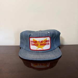 May include: A blue denim baseball cap featuring a rectangular white patch on the front. The patch has a red border and contains the yellow graphic of two crossed tobacco leaves with the red text ONTARIO FLUE-CURED TOBACCO PRODUCER.