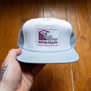 May include: A pale blue and white trucker hat with a mesh back. The hat features a rectangular patch with the text "THE ALCO GROUP WATCH US LAY!" in red and white.
