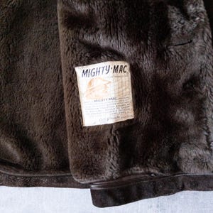 K&ouml;nnte beinhalten: Nahaufnahme des Innenraums einer braunen Mighty-Mac-Jacke. Das weiche, dunkelbraune Futter kontrastiert mit dem Etikett, das den Markennamen und eine Grafik eines Hutes zeigt. Das Etikett enth&auml;lt Details &uuml;ber die Konstruktion der Jacke.