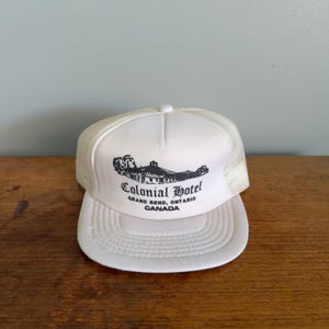 Op de afbeelding: Een witte truckerpet met een gebogen klep en mesh achterkant. Het voorpaneel toont een zwarte afbeelding van een gebouw en de woorden "Colonial Hotel, Grand Bend, Ontario, CANADA."